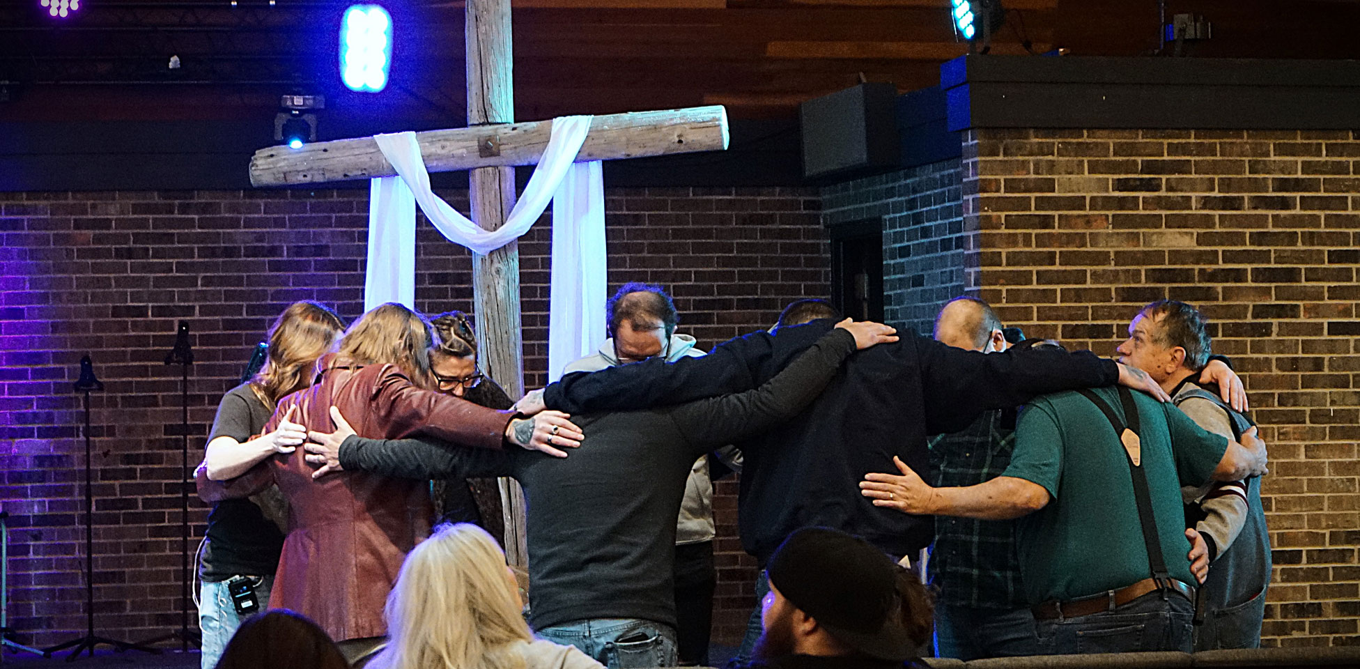 a group of people in a circle with arms around each other praying under a cross
